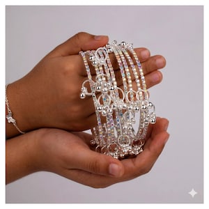 May include: A set of silver-toned bangles featuring rows of iridescent, multi-coloured rhinestones and small dangling silver charms. The bracelets have a reflective metallic finish and are designed with a thin, circular band structure.