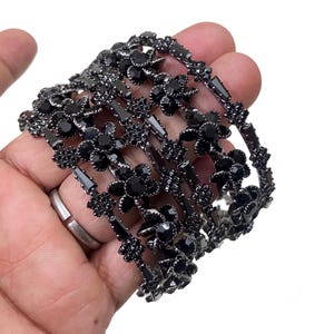 Könnte beinhalten: Ein breites Armband mit dunkelgrauem Metall-Finish und komplexen Blumenmustern. Die Oberfläche ist dicht besetzt mit zahlreichen kleinen, facettierten schwarzen Kristallen und größeren runden schwarzen Steinen in einem botanischen Motiv.
