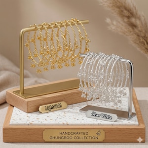 Könnte beinhalten: Gold- und silberfarbene Armreifen mit baumelnden Glöckchen werden auf einem Holz- und Marmormuster-Ständer präsentiert. Der Ständer hat einen goldfarbenen Metallrahmen und die Aufschriften "Light Gold" und "Silver White". Auf dem Sockel steht "HANDCRAFTED GHUNGROO COLLECTION".