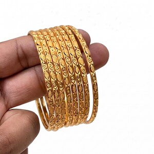 May include: A set of seven thin gold-toned bangles featuring a repeating embossed pattern of alternating circles and elongated oval shapes. Each bangle has a polished metallic finish with textured details along the outer surface of the circular band.