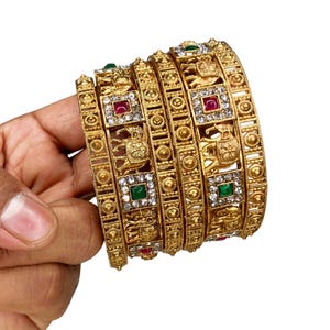 May include: A set of gold-colored bangles with intricate designs. Each bangle features square-shaped accents with red, green, and clear gemstones. The bangles have detailed patterns and include animal motifs.