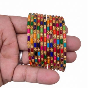 Peut inclure: Un ensemble de bracelets joncs empilables présentant des segments rectangulaires alternés dans des couleurs vives comme le rouge, le bleu, le vert, l'orange et le violet, séparés par de petits accents métalliques dorés circulaires.