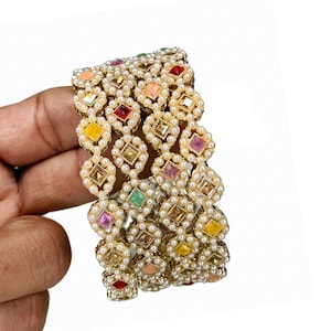 May include: A set of gold-toned bangles with a repeating pattern of small, white faux pearls and colorful square gemstones. The gemstones are in shades of red, yellow, pink, green, and orange. The bangles are held in a hand.