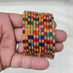 May include: A set of ten gold-toned bangles featuring alternating rectangular multi-colored enamel segments and small circular gold-toned floral accents. The bangles are arranged in a stack, displaying a vibrant array of red, blue, green, orange, and pink colors.