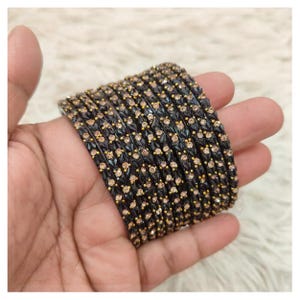 May include: A stack of black bangles with gold accents and small, light-colored gemstones. The bangles are held in a hand, showcasing their intricate design and the contrast between the dark base and the sparkling embellishments.