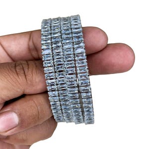 May include: A stack of five silver bracelets, each adorned with rectangular, clear crystals. The bracelets are held in a hand against a white background. The crystals reflect light, creating a sparkling effect.