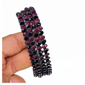 May include: A set of three circular bangles featuring alternating rows of dark black metal settings and vibrant magenta circular faceted stones. The bangles are constructed with a repeating pattern of dark metal and bright pink crystals throughout.
