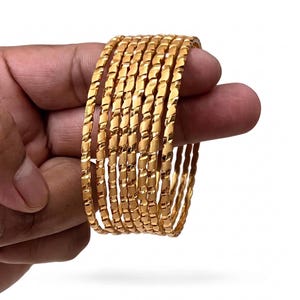 Peut inclure: Un ensemble de sept bracelets joncs de couleur dorée présentant un motif torsadé et texturé répété sur la surface extérieure. Chaque bracelet possède une finition polie qui reflète la lumière, créant un aspect métallique uniforme.