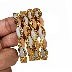 May include: Four gold bangles with a twisted design, featuring sections of silver and rose gold. The bangles have a textured pattern and are adorned with small black accents. The jewelry is held against a white background.