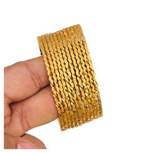 May include: A collection of gold-coloured bangles with a textured, twisted design. The bangles are held in a hand, showcasing their intricate pattern and shiny finish. The jewellery is displayed against a white background.