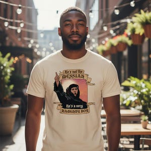 May include: Cream-coloured t-shirt with a graphic of a person in a black robe pointing upwards, with the text "He's not the MESSIAH" and "He's a very NAUGHTY BOY". The shirt is worn by a person.