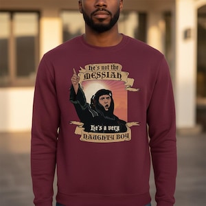 May include: A burgundy sweatshirt featuring a graphic of a man in a black robe, pointing upwards. The text on the sweatshirt reads "he's not the MESSIAH" above the image and "he's a very NAUGHTY BOY" below.
