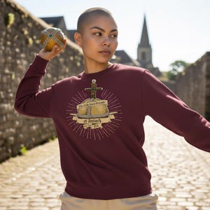 May include: Burgundy crew neck sweatshirt with a graphic of a crown and cross, and the text "The Holy Hand Grenade of Antioch". The person in the image is holding a decorative ball. The background includes a stone wall and a church spire.