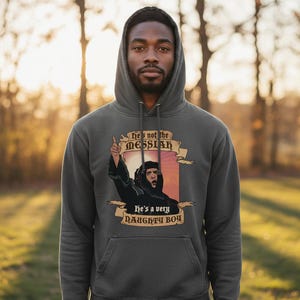 May include: Charcoal grey hoodie with a graphic of a person in a black robe, with the text "He's not the MESSIAH, he's a very NAUGHTY BOY." The design is in a banner style with a pink and yellow background.