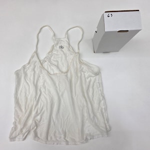 May include: A white racerback tank top with thin shoulder straps and a scoop neckline. The garment features a small grey logo on the upper back area. A white rectangular cardboard box with the handwritten number 63 is positioned nearby.