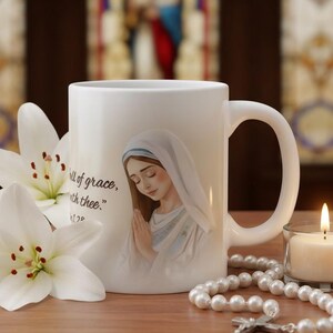 Op de afbeelding: Witte keramische mok met een illustratie van een vrouw in een witte sluier en de tekst "full of grace, the Lord is with thee." Witte lelies, een brandende kaars en een parelrozenkrans staan ook in de afbeelding.