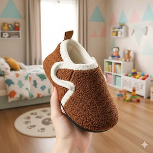 May include: Brown, textured slipper with a white trim and a small brown loop. The interior is white and fluffy. The slipper is held in a hand, with a blurred background of a child's bedroom.