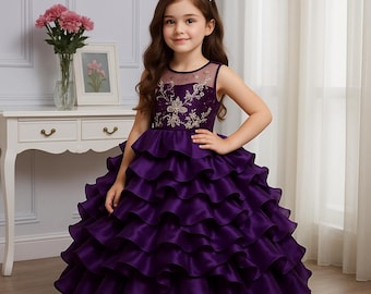 May include: A young girl wearing a formal purple dress with layered ruffles. The sleeveless bodice features embroidered gold detailing. She wears a silver tiara. The dress is suitable for a special occasion.