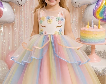 May include: A young girl in a rainbow-colored unicorn-themed dress. The dress features a white bodice with floral accents and a tulle skirt in pastel shades. Two unicorn balloons and a rainbow cake are in the background.