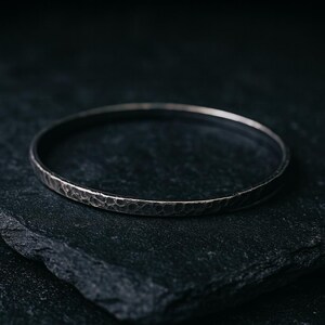 May include: A silver-colored bangle bracelet with a hammered texture. The bracelet is circular and sits on a dark, textured surface. The metal has a slightly tarnished appearance.