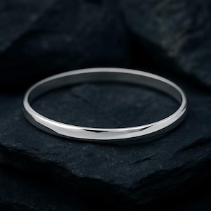 Könnte beinhalten: Ein poliertes silbernes Armreif-Armband. Das Armband hat ein einfaches, abgerundetes Design mit einer glatten, reflektierenden Oberfläche. Der Schmuck wird auf einer dunklen, strukturierten Oberfläche präsentiert.