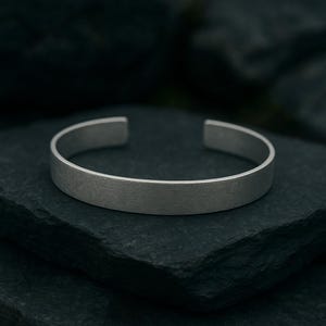 May include: A silver cuff bracelet with a brushed metal finish. The open design allows for easy wear. The bracelet is a simple, minimalist accessory.