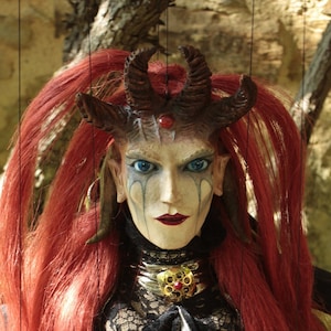 May include: A red-haired puppet with horns, wearing a black lace dress with gold accents. The puppet has blue eyes and a painted face.
