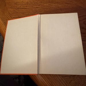 K&ouml;nnte beinhalten: Ein offenes Buch mit leeren Seiten. Das Buch hat einen leuchtend orangefarbenen Buchr&uuml;cken und Kanten. Die Seiten sind hellbraun. Das Buch liegt auf einer Holzoberfl&auml;che.