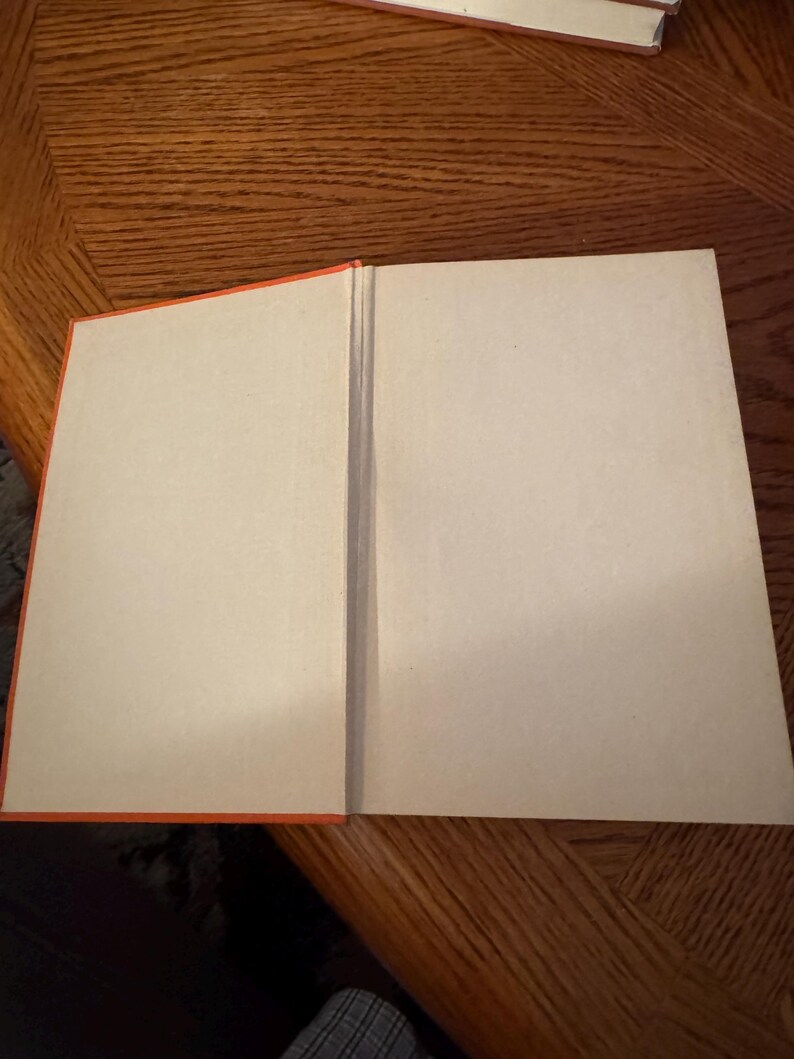 K&ouml;nnte beinhalten: Ein offenes Buch mit leeren Seiten. Das Buch hat einen leuchtend orangefarbenen Buchr&uuml;cken und Kanten. Die Seiten sind hellbraun. Das Buch liegt auf einer Holzoberfl&auml;che.