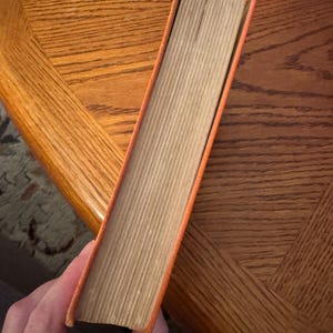 K&ouml;nnte beinhalten: Ein orangefarbenes Buch mit einem abgenutzten R&uuml;cken und gealterten Seiten. Die Buchkanten sind orange und die Seiten hellbraun. Das Buch wird schr&auml;g gehalten, wodurch die Seite der Seiten sichtbar wird.