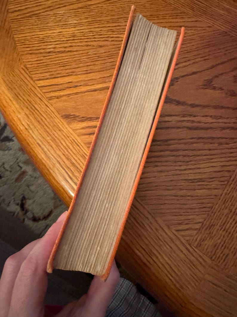 K&ouml;nnte beinhalten: Ein orangefarbenes Buch mit einem abgenutzten R&uuml;cken und gealterten Seiten. Die Buchkanten sind orange und die Seiten hellbraun. Das Buch wird schr&auml;g gehalten, wodurch die Seite der Seiten sichtbar wird.