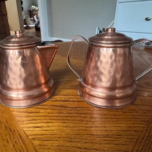 May include: Two copper teapots with hammered textures sit on a wooden table. The teapot on the left has a spout and a handle, while the one on the right has a rounded shape with a lid and a handle. Both are vintage and have a shiny finish.