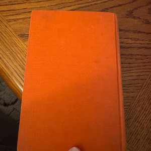 K&ouml;nnte beinhalten: Ein Buch mit leuchtend orangefarbenem Einband. Das Buch wird von einer Hand gehalten, wobei der Einband nach oben zeigt. Der Buchr&uuml;cken ist sichtbar, und der Einband hat eine strukturierte Oberfl&auml;che. Der Hintergrund ist ein Holztisch.