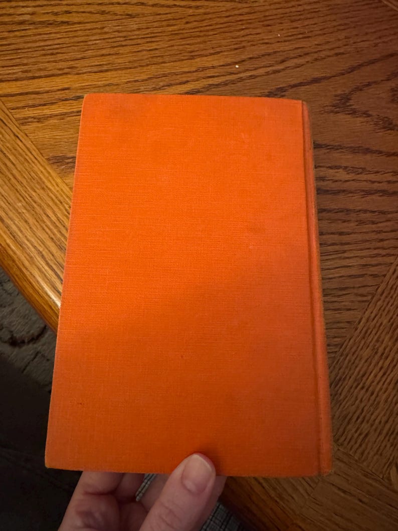 K&ouml;nnte beinhalten: Ein Buch mit leuchtend orangefarbenem Einband. Das Buch wird von einer Hand gehalten, wobei der Einband nach oben zeigt. Der Buchr&uuml;cken ist sichtbar, und der Einband hat eine strukturierte Oberfl&auml;che. Der Hintergrund ist ein Holztisch.