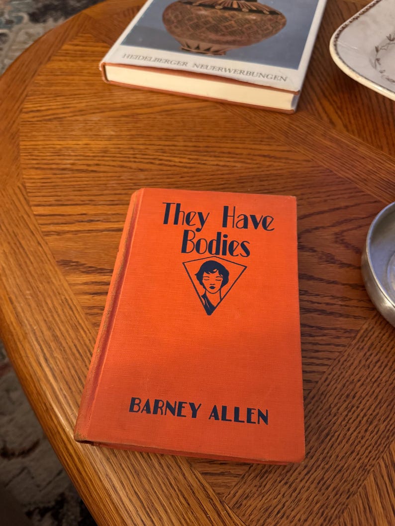 K&ouml;nnte beinhalten: Ein orangefarbenes Buch mit dem Titel "They Have Bodies" von Barney Allen. Das Cover zeigt eine blau-wei&szlig;e Illustration eines Frauengesichts in einem Dreieck. Das Buch liegt auf einem Holztisch, im Hintergrund weitere B&uuml;cher und Gegenst&auml;nde.