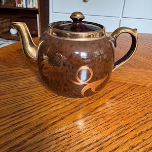 May include: A round, dark brown ceramic teapot featuring a glossy finish, a matching lid with a gold-toned knob, and a gold-toned spout and handle. The body of the teapot is decorated with a stylized, metallic gold circular and leaf-like pattern.