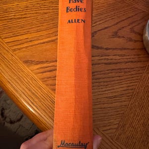 K&ouml;nnte beinhalten: Ein Vintage-Buch mit dem Titel "They Have Bodies" von Allen. Das Buch hat einen orangefarbenen Einband mit dem Titel und dem Namen des Autors in Schwarz. Der Name des Verlags, Macaulay, ist unten aufgedruckt. Das Buch wird vertikal gehalten.