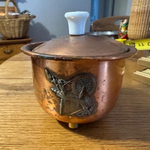 May include: A copper-colored container with a lid and a white knob. The container has a decorative emblem on the side. The container is sitting on a wooden table. The container is likely used for storage or display.