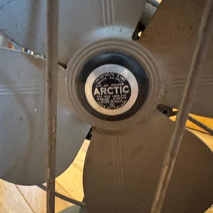 May include: Vintage Arctic fan with four blades and a protective cage. The fan blades are a muted gray color, and the center features a circular emblem with the word "ARCTIC". The fan is set against a wooden background.
