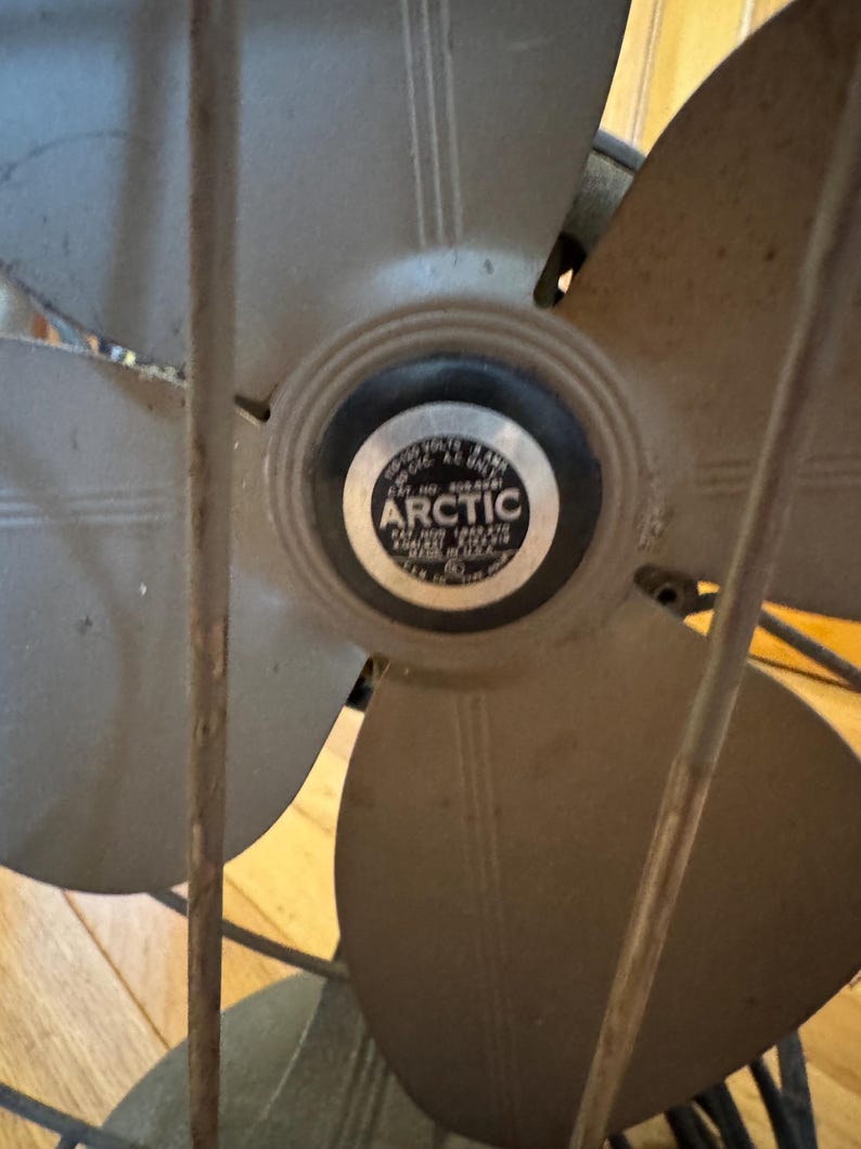 May include: Vintage Arctic fan with four blades and a protective cage. The fan blades are a muted gray color, and the center features a circular emblem with the word "ARCTIC". The fan is set against a wooden background.