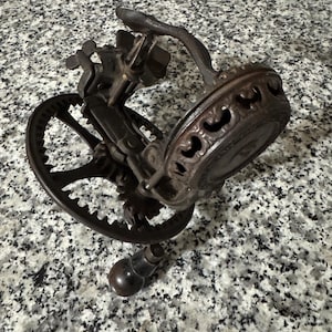 May include: An antique, cast iron hand-cranked egg beater. The beater has a dark brown finish, with a gear and handle. The beater's design includes decorative scrollwork around the base. The beater is a vintage kitchen tool.