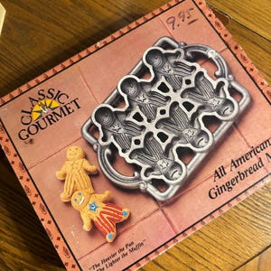 May include: A Classic Gourmet gingerbread man pan with a silver finish, designed to bake multiple gingerbread men. The box features the text "All American Gingerbread Men" and two gingerbread men cookies.