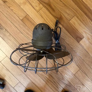 May include: An antique, bronze-colored electric fan with a cage-like design. The fan has three blades and a rounded motor housing with ventilation holes. A black power cord extends from the back. The fan sits on a wooden floor.