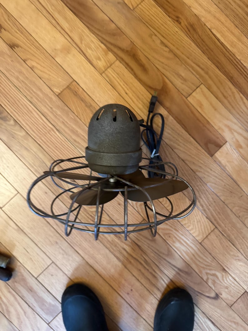 May include: An antique, bronze-colored electric fan with a cage-like design. The fan has three blades and a rounded motor housing with ventilation holes. A black power cord extends from the back. The fan sits on a wooden floor.