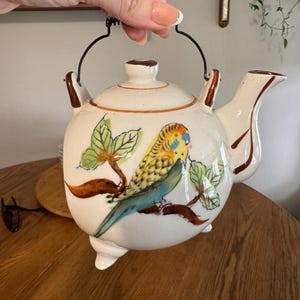 May include: A ceramic teapot with a colorful parakeet design. The white teapot features a black handle and brown accents. The parakeet is depicted with yellow, blue, and green feathers, perched on a brown branch with green leaves. The teapot has a small spout and three feet.