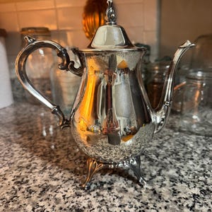 May include: A silver-colored teapot with a curved handle and spout, resting on three decorative feet. The teapot has a lid with a finial and reflects the surrounding environment. The teapot is likely used for serving tea.