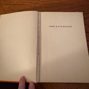 K&ouml;nnte beinhalten: Ein offenes Buch mit dem Text "THEY HAVE BODIES" auf der rechten Seite. Die Seiten sind cremefarben, und der Buchr&uuml;cken ist sichtbar. Der Buchrand ist orange. Das Buch liegt auf einer Holzoberfl&auml;che.