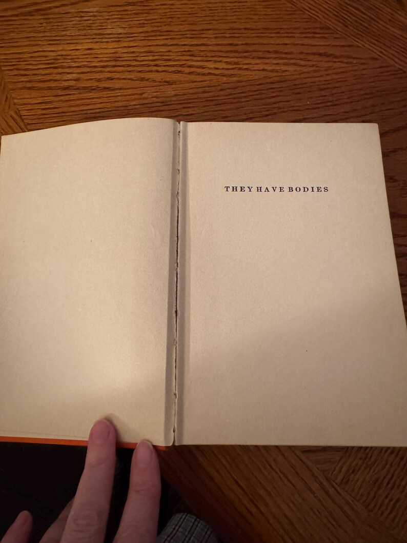 K&ouml;nnte beinhalten: Ein offenes Buch mit dem Text "THEY HAVE BODIES" auf der rechten Seite. Die Seiten sind cremefarben, und der Buchr&uuml;cken ist sichtbar. Der Buchrand ist orange. Das Buch liegt auf einer Holzoberfl&auml;che.