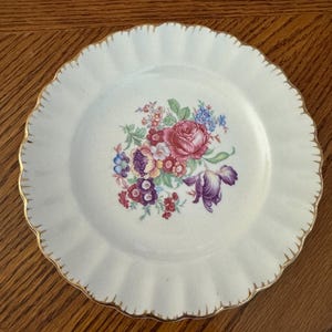 May include: A white ceramic plate with a scalloped edge and gold trim. The plate features a floral bouquet design in shades of pink, purple, blue, and yellow. The plate is set on a wooden surface.