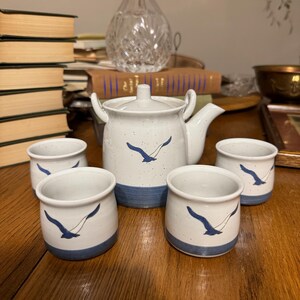 May include: A white ceramic tea set with a teapot and four cups. Each piece features a blue seagull design and a blue band at the base. The teapot has a lid and a spout. The set is on a wooden surface.